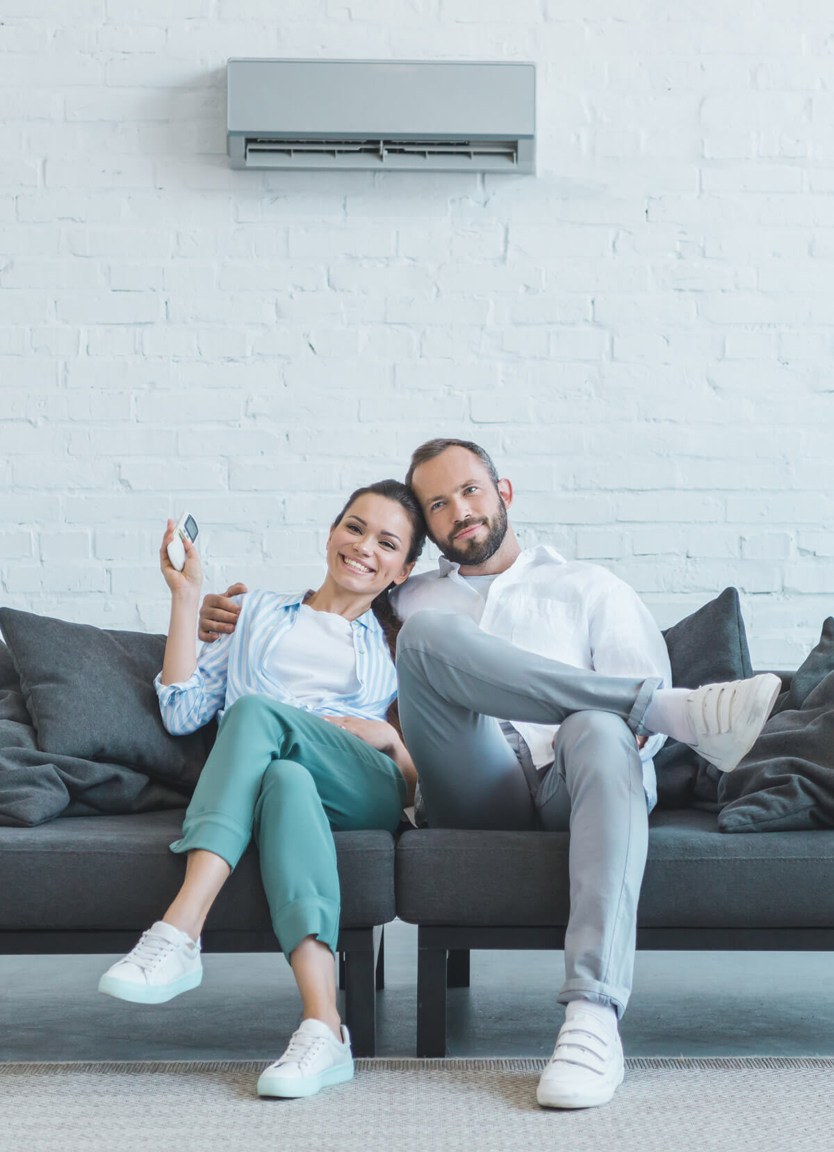 A woman and a man sit on a gray sofa. The woman is controlling a wall mounted mini-split HVAC unit with a remote.