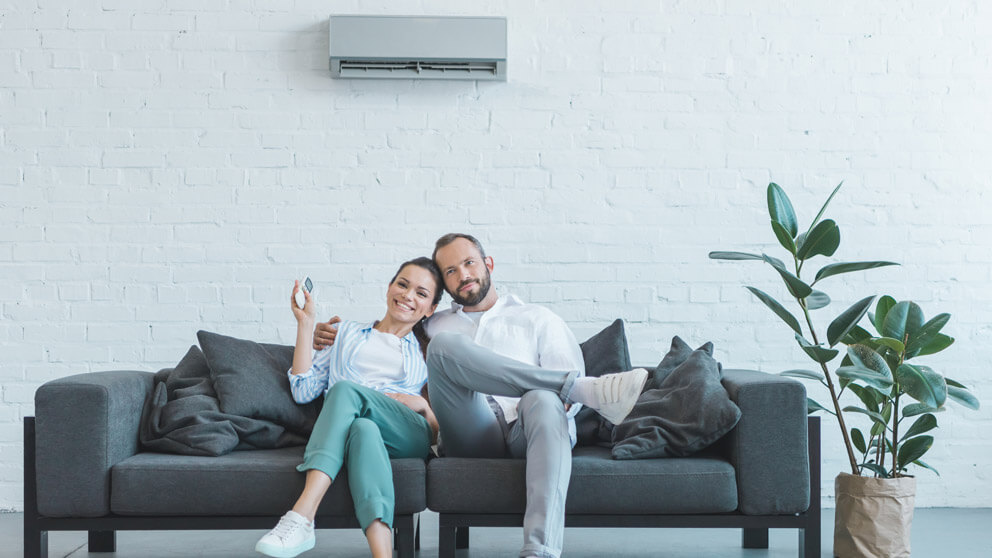 A woman and a man sit on a gray sofa. The woman is controlling a wall mounted mini-split HVAC unit with a remote.