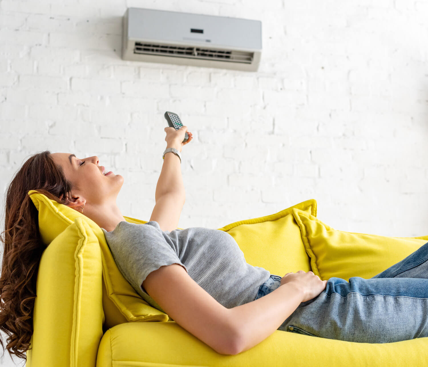 A smiling woman relaxes on a couch. She is pointing a remote at a mini-split air conditioner.